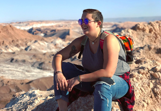 young person with short hair, sunglasses and a backpack sitting on a rocky mountain